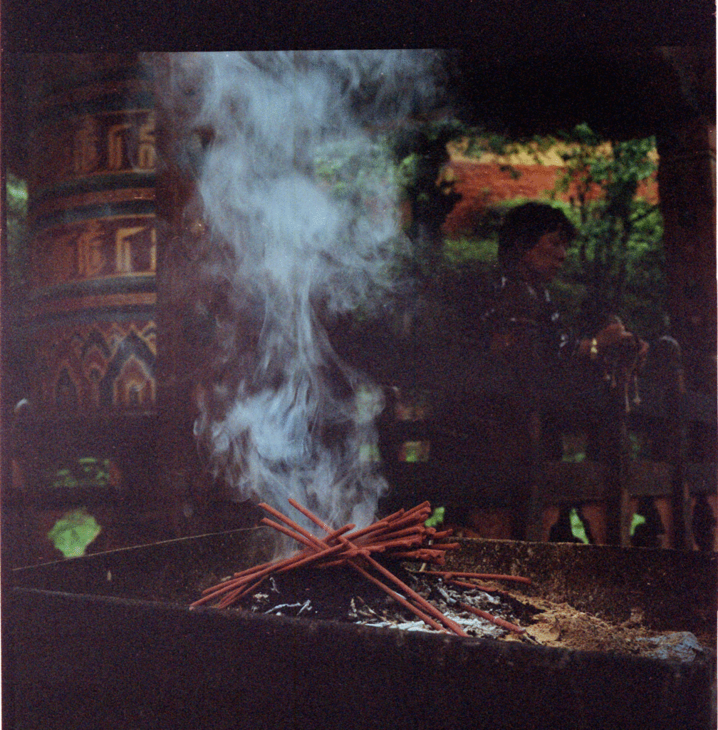 Incense piled up in a smoke offering at the entrance to Dechenphug temple, in the shadowy background visitors turn a large prayer wheel