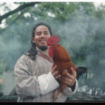 Wang wears a gho and holds a rooster up to the camera