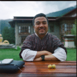 Yeshi Lhendup wears a gho and sits at an outdoor table in front of the Department of Culture
