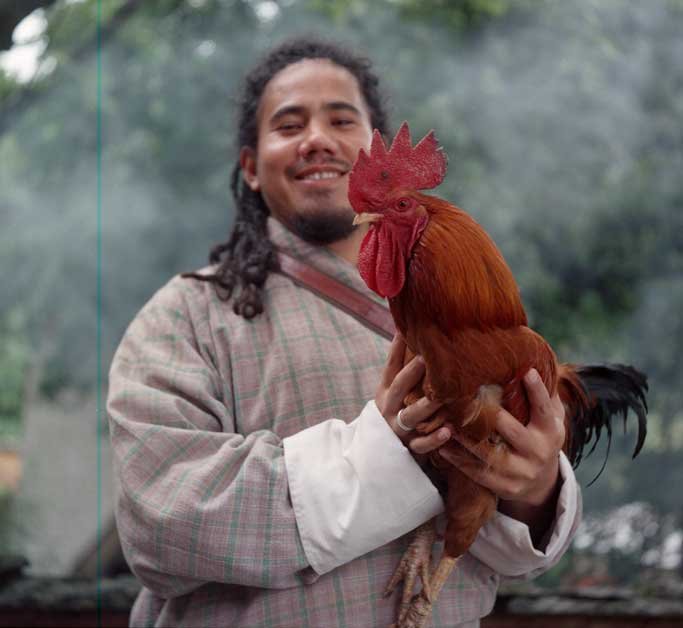 2025June19_Thimphu_Y_KP800_Denchenphug_DfT_Wang Wang wears a gho and holds up a rooster at a lhakhang
