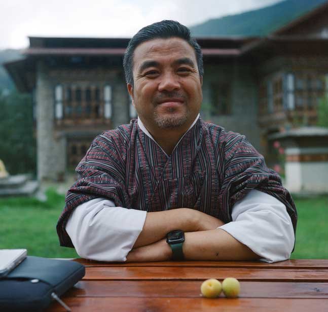 25June224_Thimphu_Y_KPortra800_-7_NLAB_YeshiLhendup Yeshi crosses his arms on a table and smiles widely. He's at the Department of culture with fruit in front of him